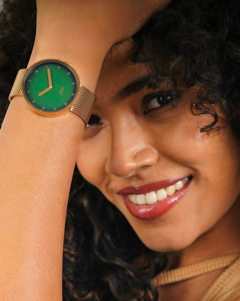 Woman wearing a watch with a green face and gold band, smiling.